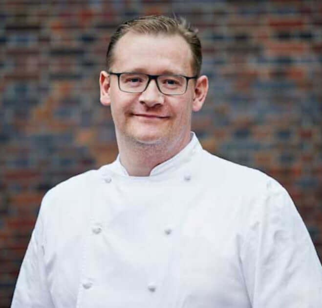 Christian Krüger in Kochoutfit, lächelnd in die Kamera schauend. Sein freundliches Lächeln und seine professionelle Ausstrahlung machen ihn zu einem sympathischen Koch im Portrait